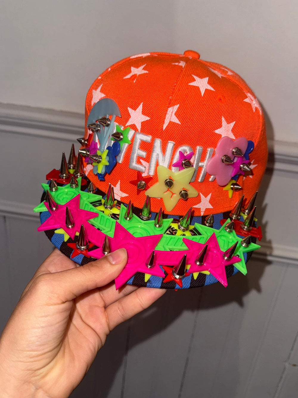 “An orange baseball cap decorated with white stars and the word ‘GIVENCHY’ embroidered in white letters. The brim and front are adorned with colorful 3D embellishments, including bright pink and green star shapes, multicolored flower designs, and metallic silver spikes, creating a bold and playful aesthetic. The cap is held in a hand for display.”