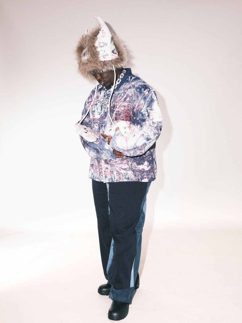 “Person wearing a vibrant, artistic outfit featuring a textured, multicolored jacket with abstract patterns, paired with dark pants accented by light denim panels. They are also wearing a unique pointed hat with a fur trim, adorned with illustrations and text, and black shoes. The individual is posed in a minimalistic studio setting with a neutral backdrop, looking downward.”