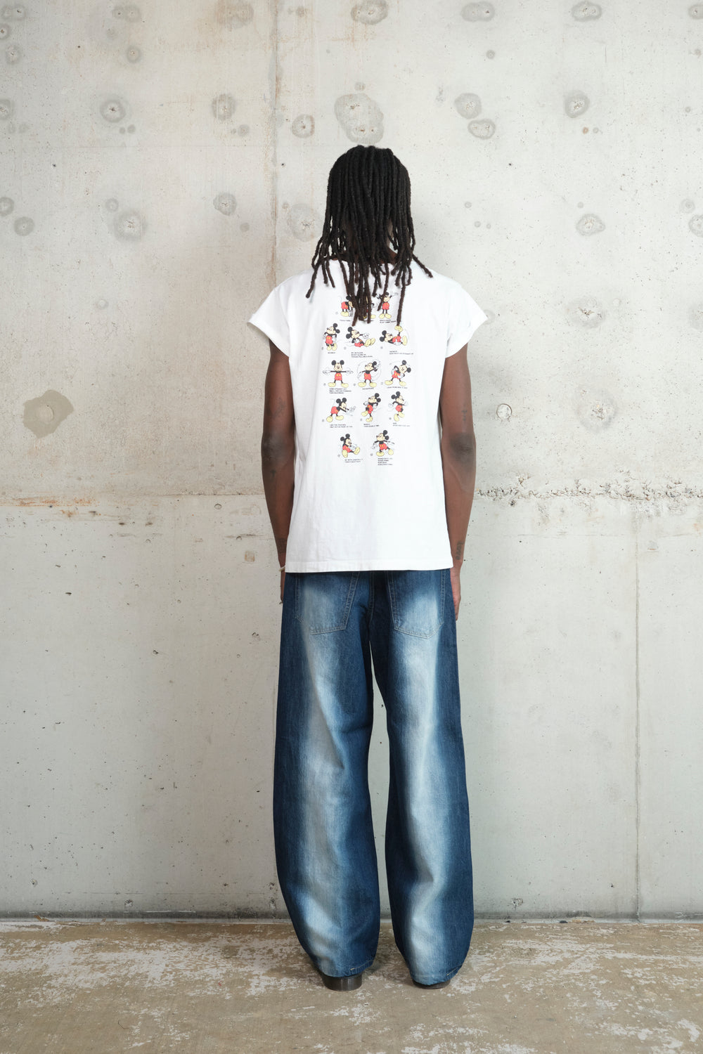 A person with long dreadlocks stands with their back to the camera, wearing a white Gatto T-shirt with a small graphic print on the upper back featuring cartoon characters in various fitness poses. The shirt has rolled-up sleeves and a relaxed fit. They are also wearing wide-leg, faded denim jeans with a vintage wash. The backdrop is a raw concrete wall, adding to the minimal, industrial aesthetic.