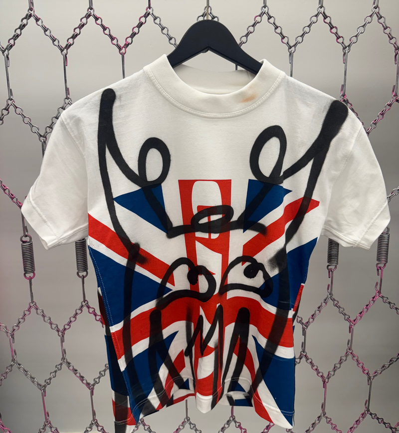 White t-shirt with British flag design and black graffiti-style text on a textured wall background.