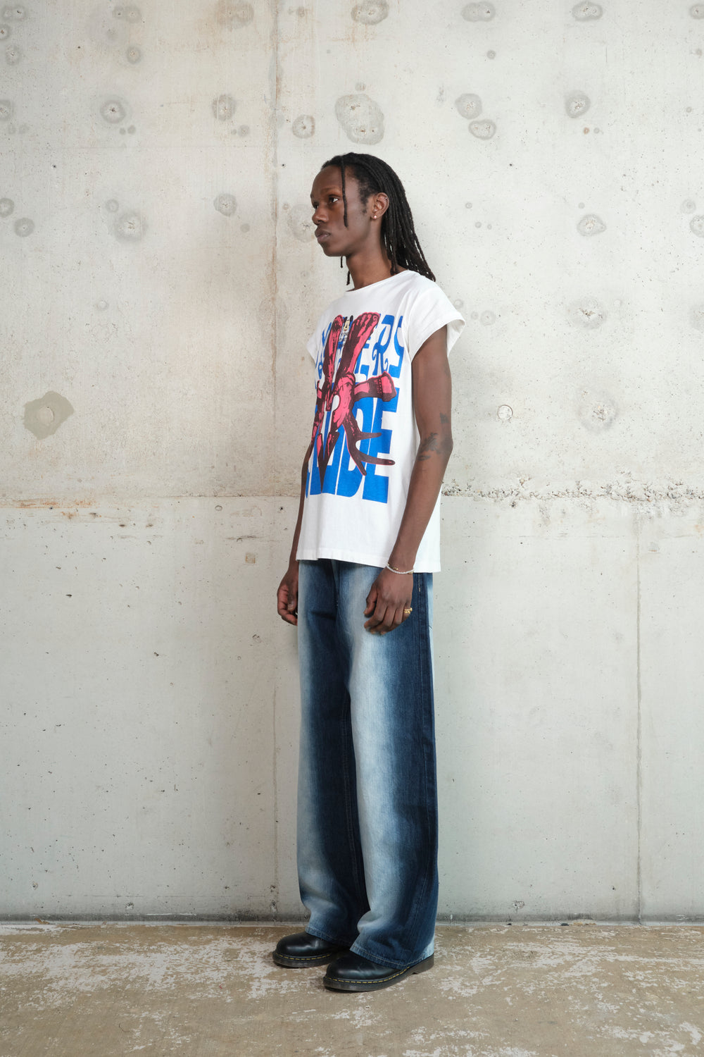 A person with long dreadlocks wears a white Gatto T-shirt with a bold red and blue screenprint, featuring skeletal hands gripping wrenches over block lettering. The shirt has rolled-up sleeves and a relaxed fit. They are also wearing wide-leg, faded denim jeans and black leather shoes. Standing in a neutral pose against a raw concrete wall, they gaze to the side with a composed expression.
