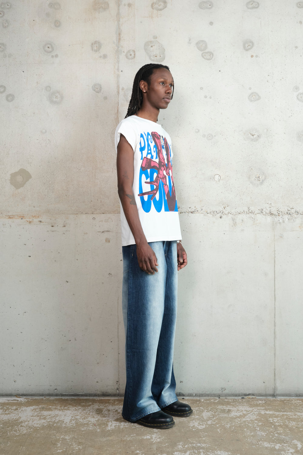 A person with long dreadlocks stands in profile against a raw concrete wall, wearing a white Gatto T-shirt with a bold red and blue screenprint design on the front. The relaxed-fit shirt has rolled-up sleeves. They pair it with wide-leg, faded denim jeans and black shoes, creating a casual yet striking silhouette.