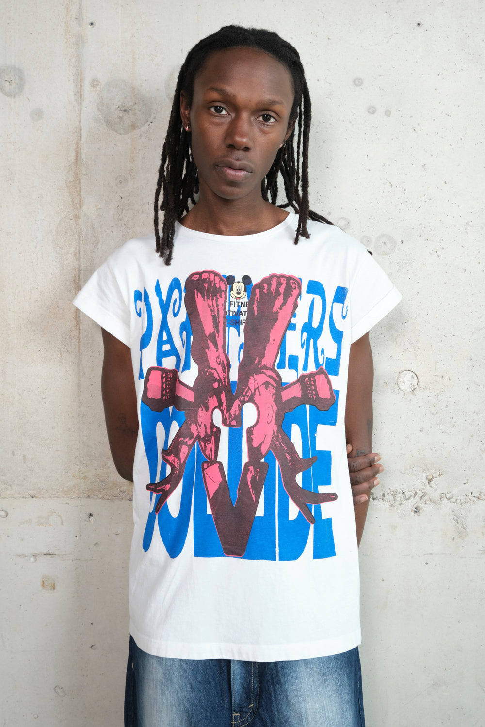 A person with long dreadlocks wears a white Gatto T-shirt with a bold screenprint design. The print features red skeletal hands gripping wrenches over large blue block letters spelling PARTNERS COLLIDE. The T-shirt has rolled-up sleeves and a relaxed fit, paired with light-wash denim jeans. The subject stands against a raw concrete wall, gazing directly at the camera with a composed expression.