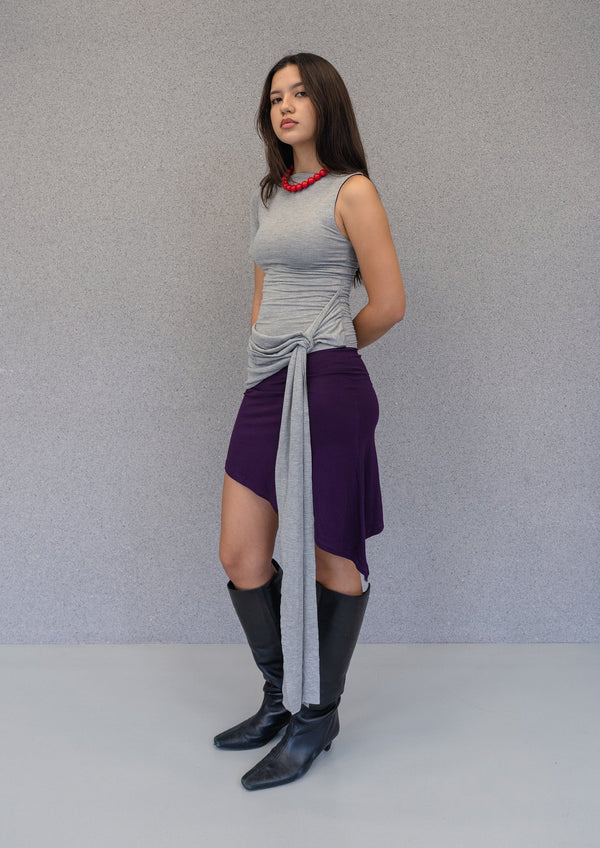 Front view of grey and purple asymmetrical mini dress with draped waist detail and one-shoulder sleeve worn with black knee-high boots.