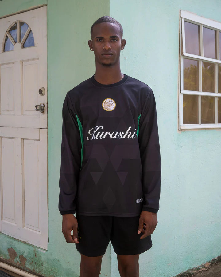 A young man with short hair and a lean build stands in front of a pastel green house with a white door and a window. He is wearing a black long-sleeve sports jersey with subtle geometric patterns, green accents on the sides, and the word ‘Kurashi’ written in white cursive across the chest. A gold emblem is positioned on the upper left side of the jersey. He pairs the jersey with black athletic shorts and has a calm, neutral expression as he looks directly into the camera