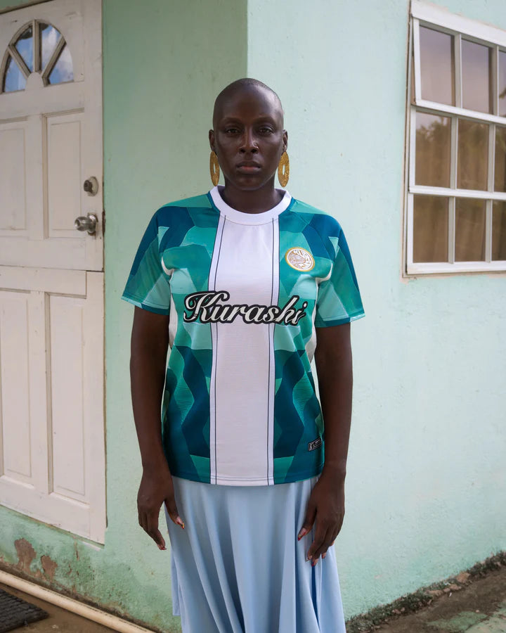 A woman with a shaved head and large gold earrings stands in front of a pastel green house with a white door and a window. She wears a green and white patterned sports jersey with the word ‘Kurashi’ printed across the chest and a flowing light blue skirt. Her expression is serious as she looks directly at the camera. The background has a textured wall and a simple residential setting