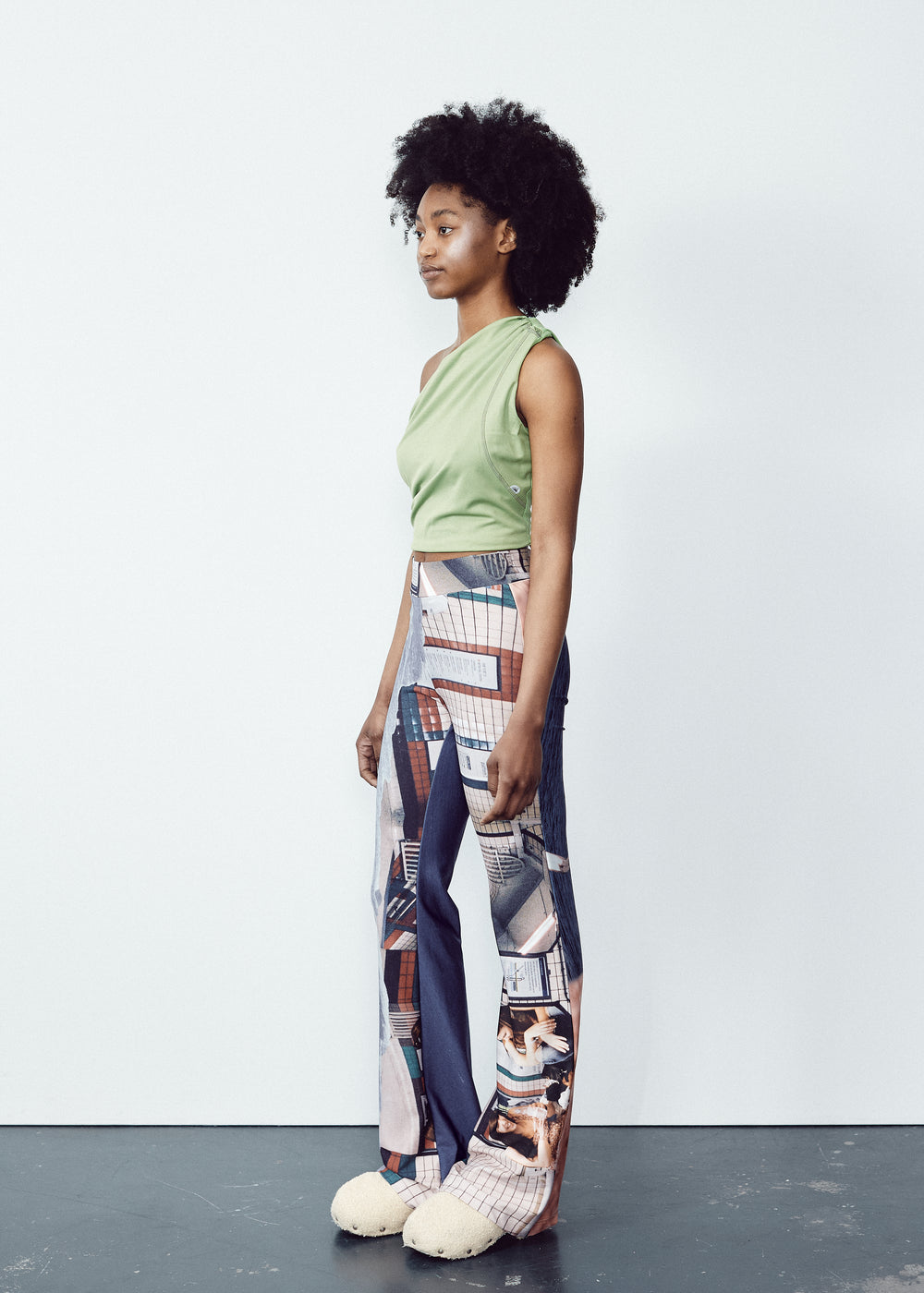 Back view of a person wearing a white long-sleeve top and colorful, abstract-print flared pants. The outfit features unique, artistic designs with a blend of blue and orange tones on the pants, contrasted by the simplicity of the white top. The model has natural, voluminous curly hair.