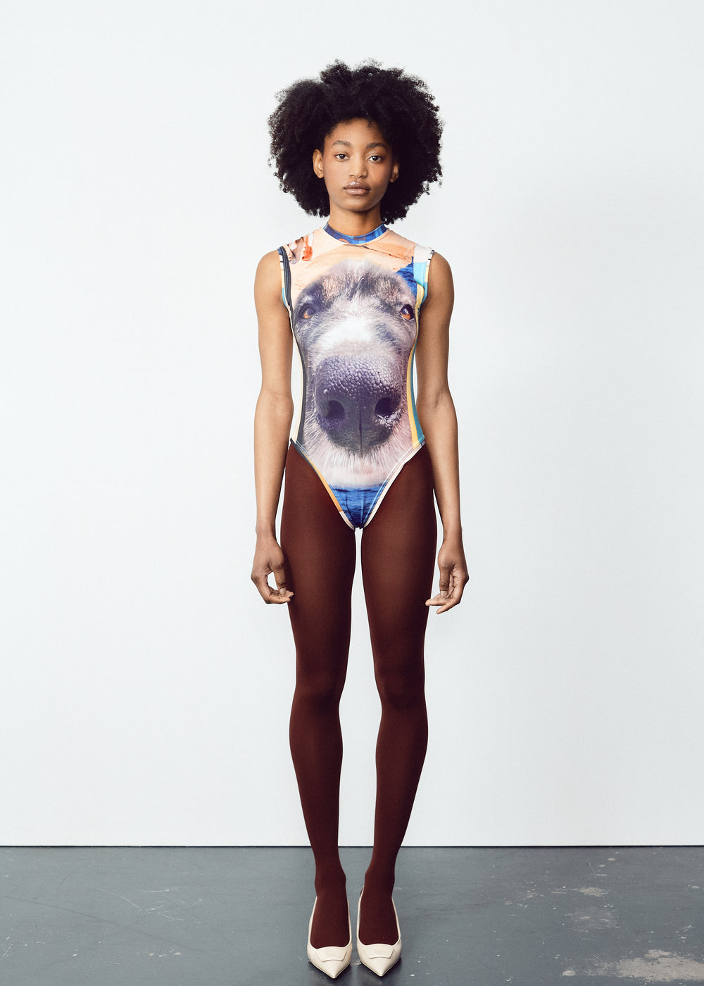 Model wearing a sleeveless bodysuit with a large, detailed dog face graphic on the front. The outfit is paired with dark brown tights and white pointed heels. The model has natural curly hair and is standing straight against a plain, light-colored backdrop