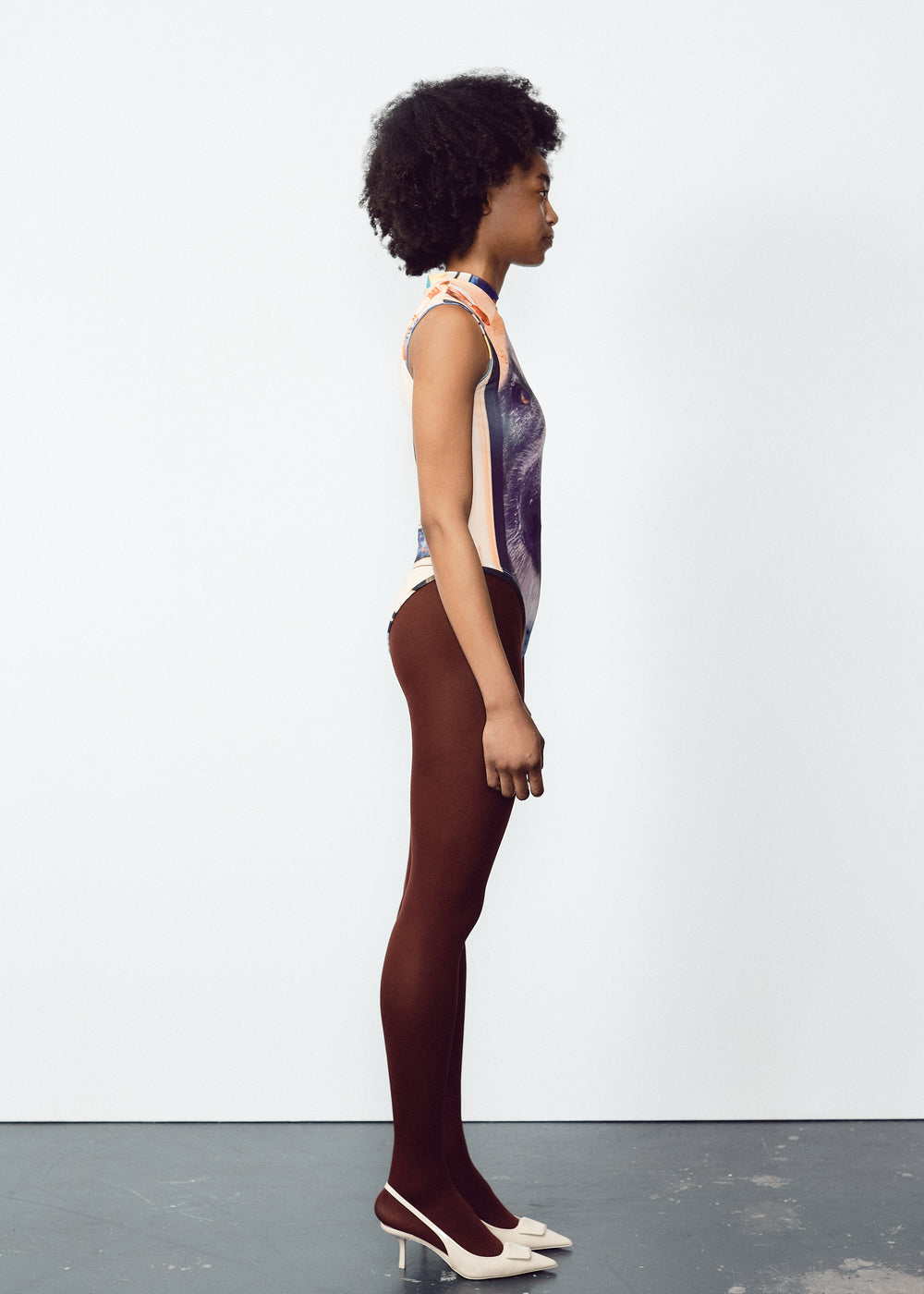 Model wearing a sleeveless bodysuit with a large, detailed dog face graphic on the front. The outfit is paired with dark brown tights and white pointed heels. The model has natural curly hair and is standing straight against a plain, light-colored backdrop