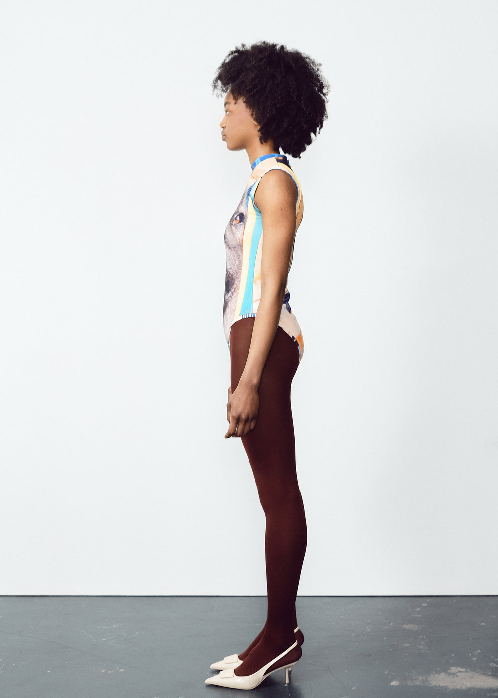 Model wearing a sleeveless bodysuit with a large, detailed dog face graphic on the front. The outfit is paired with dark brown tights and white pointed heels. The model has natural curly hair and is standing straight against a plain, light-colored backdrop