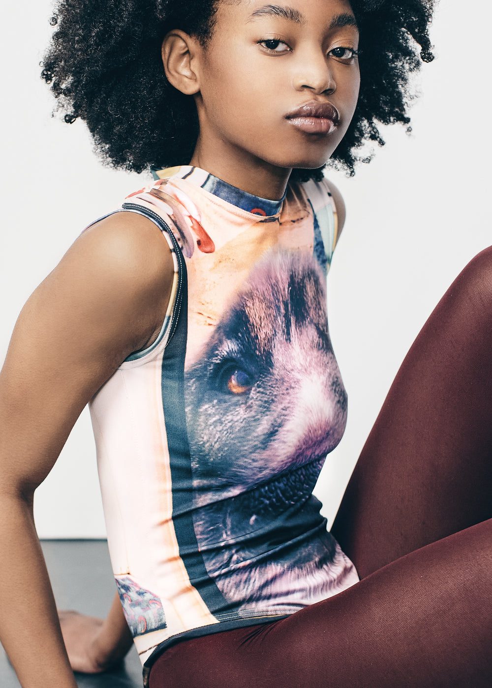 Model wearing a sleeveless bodysuit with a large, detailed dog face graphic on the front. The outfit is paired with dark brown tights and white pointed heels. The model has natural curly hair and is standing straight against a plain, light-colored backdrop