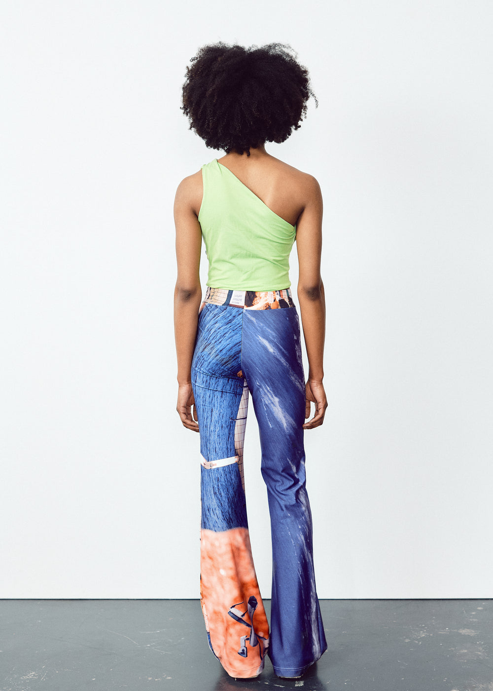 Back view of a person wearing a white long-sleeve top and colorful, abstract-print flared pants. The outfit features unique, artistic designs with a blend of blue and orange tones on the pants, contrasted by the simplicity of the white top. The model has natural, voluminous curly hair.