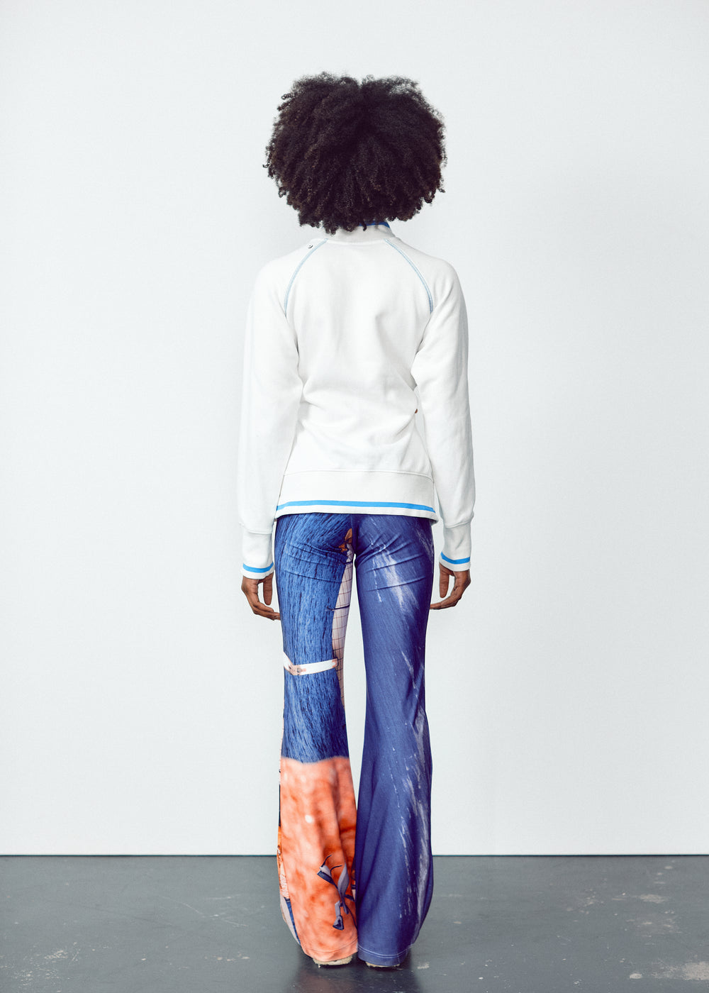 Back view of a person wearing a white long-sleeve top and colorful, abstract-print flared pants. The outfit features unique, artistic designs with a blend of blue and orange tones on the pants, contrasted by the simplicity of the white top. The model has natural, voluminous curly hair.