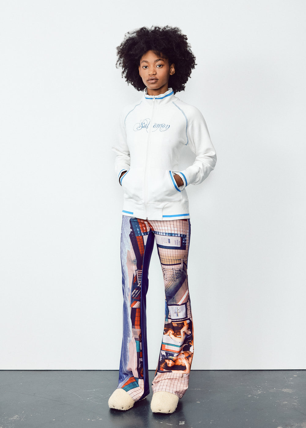 front view of a person wearing a white long-sleeve top and colorful, abstract-print flared pants. The outfit features unique, artistic designs with a blend of blue and orange tones on the pants, contrasted by the simplicity of the white top. The model has natural, voluminous curly hair.