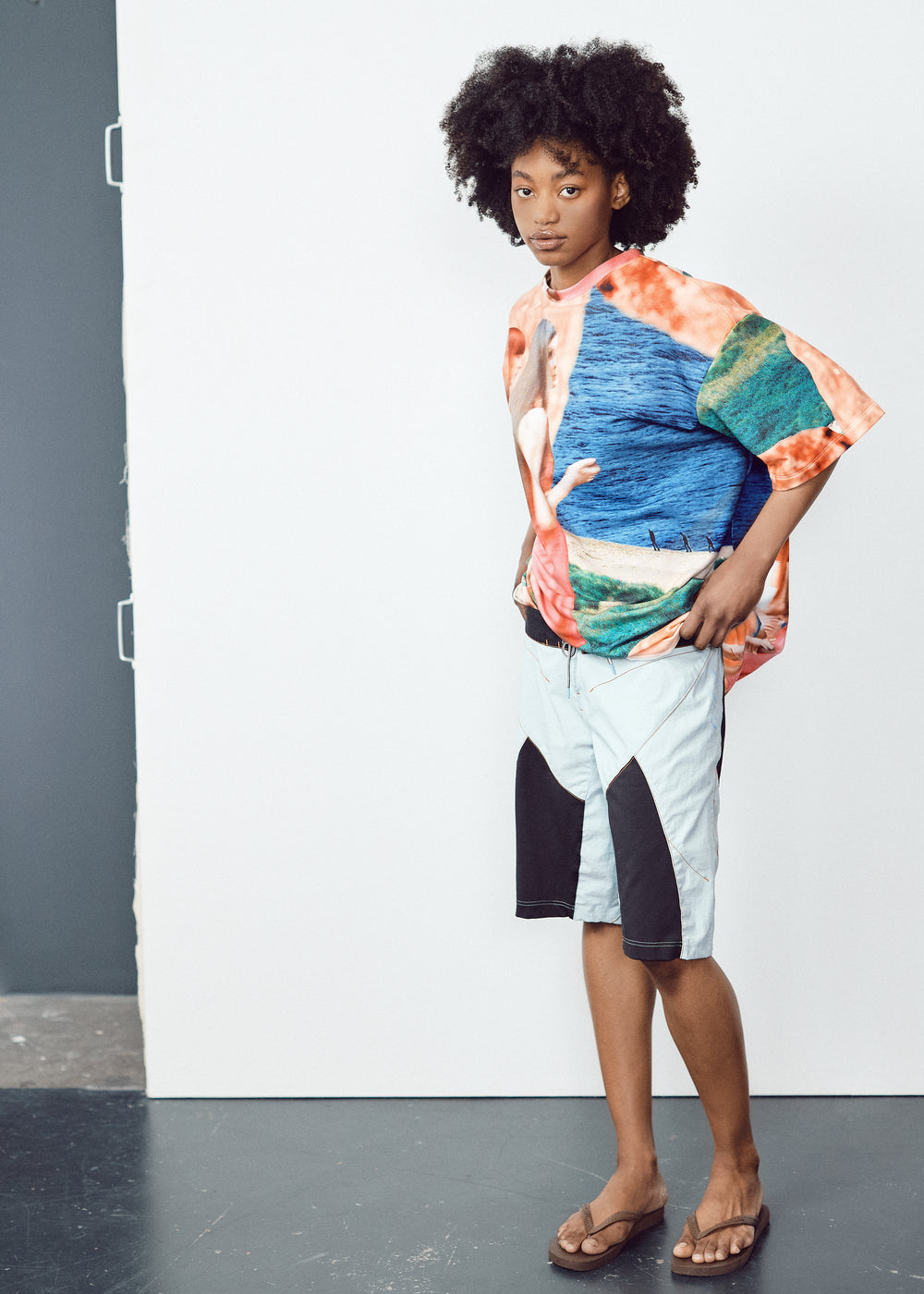 Model in a vibrant, oversized T-shirt with abstract blue, green, and orange patterns, paired with light knee-length shorts featuring black panel accents. The model has natural curly hair and wears flip-flops, standing in a relaxed pose against a minimalistic backdrop