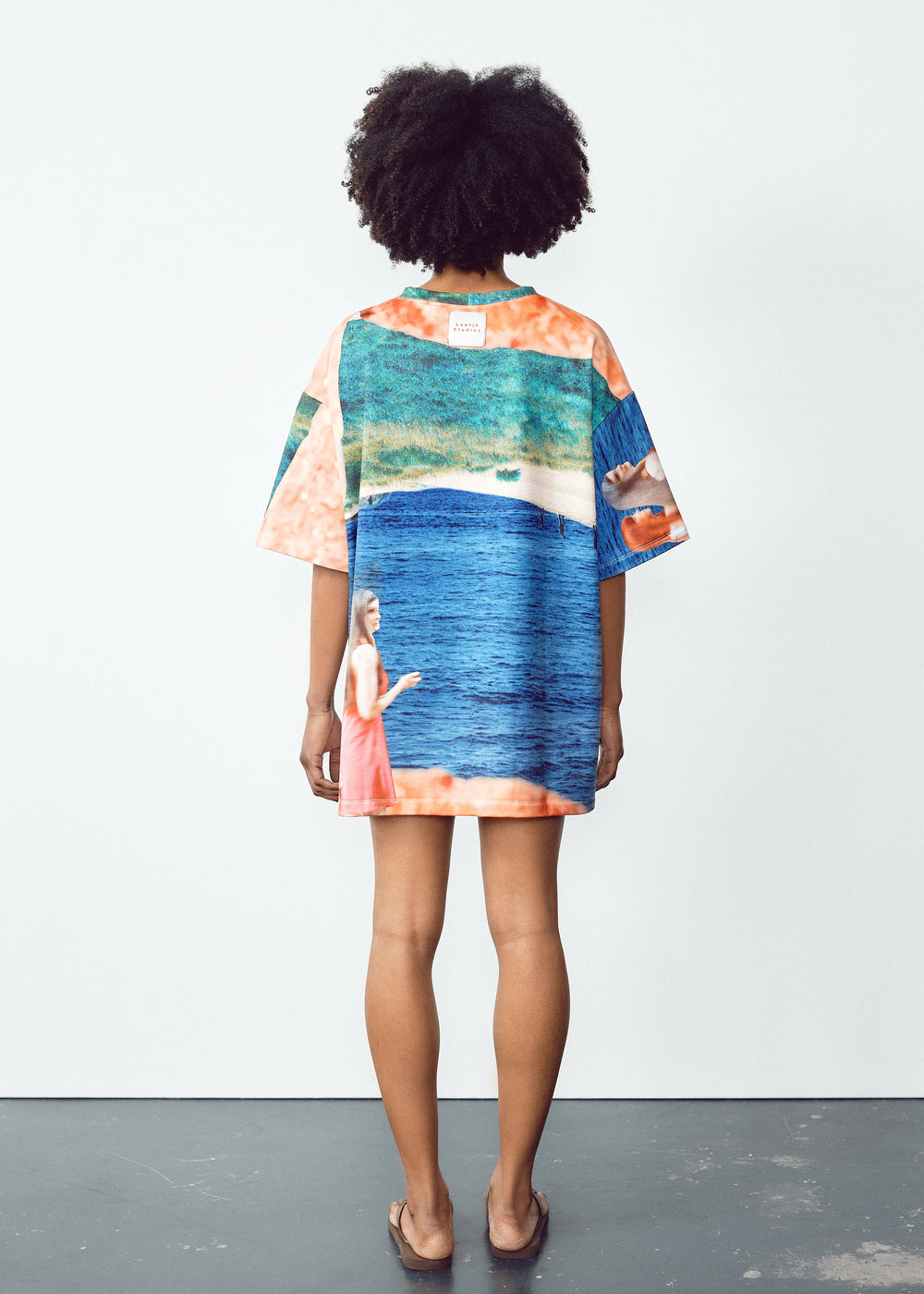 Model in a vibrant, oversized T-shirt with abstract blue, green, and orange patterns, paired with light knee-length shorts featuring black panel accents. The model has natural curly hair and wears flip-flops, standing in a relaxed pose against a minimalistic backdrop