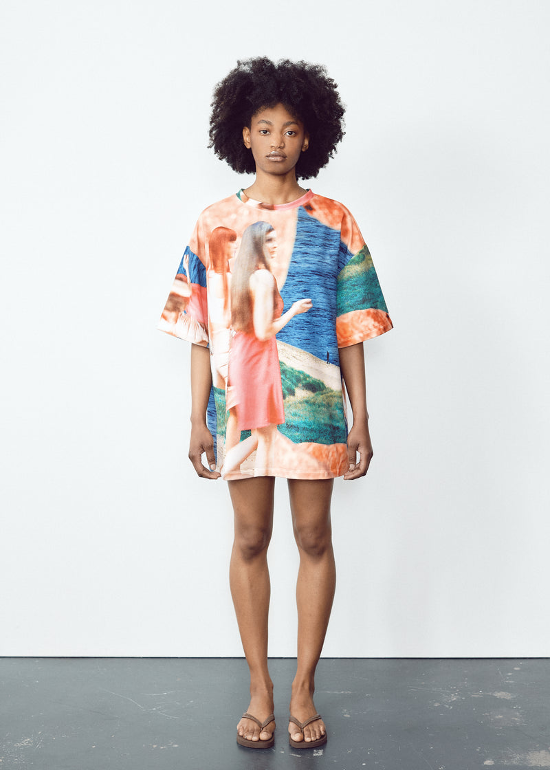 Model in a vibrant, oversized T-shirt with abstract blue, green, and orange patterns, paired with light knee-length shorts featuring black panel accents. The model has natural curly hair and wears flip-flops, standing in a relaxed pose against a minimalistic backdrop