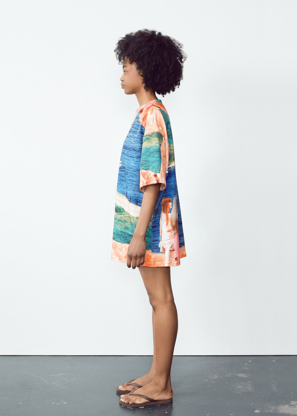 Model in a vibrant, oversized T-shirt with abstract blue, green, and orange patterns, paired with light knee-length shorts featuring black panel accents. The model has natural curly hair and wears flip-flops, standing in a relaxed pose against a minimalistic backdrop