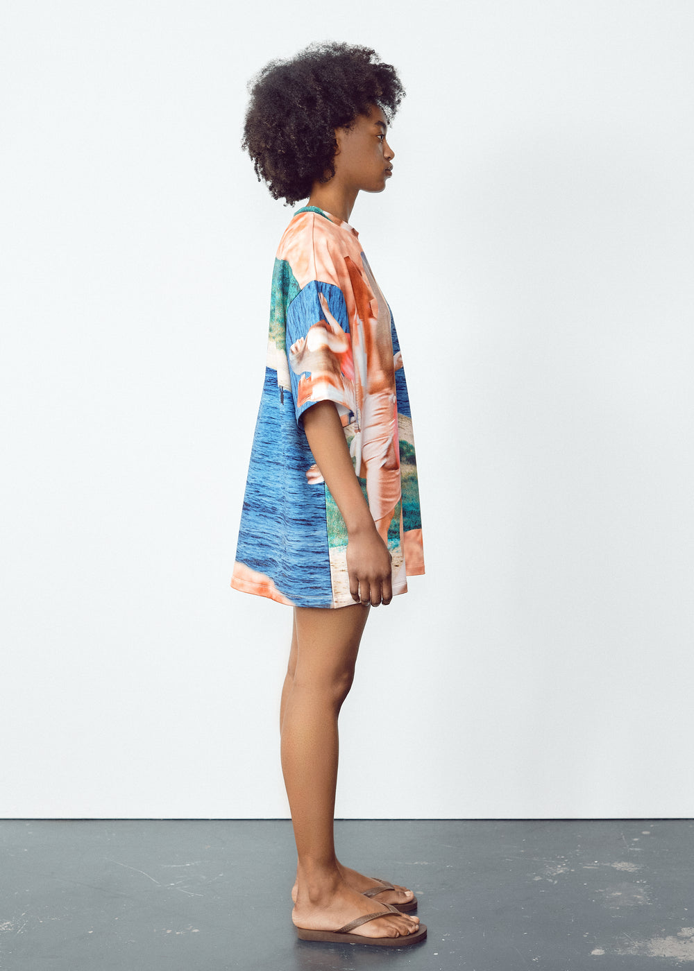 Model in a vibrant, oversized T-shirt with abstract blue, green, and orange patterns, paired with light knee-length shorts featuring black panel accents. The model has natural curly hair and wears flip-flops, standing in a relaxed pose against a minimalistic backdrop