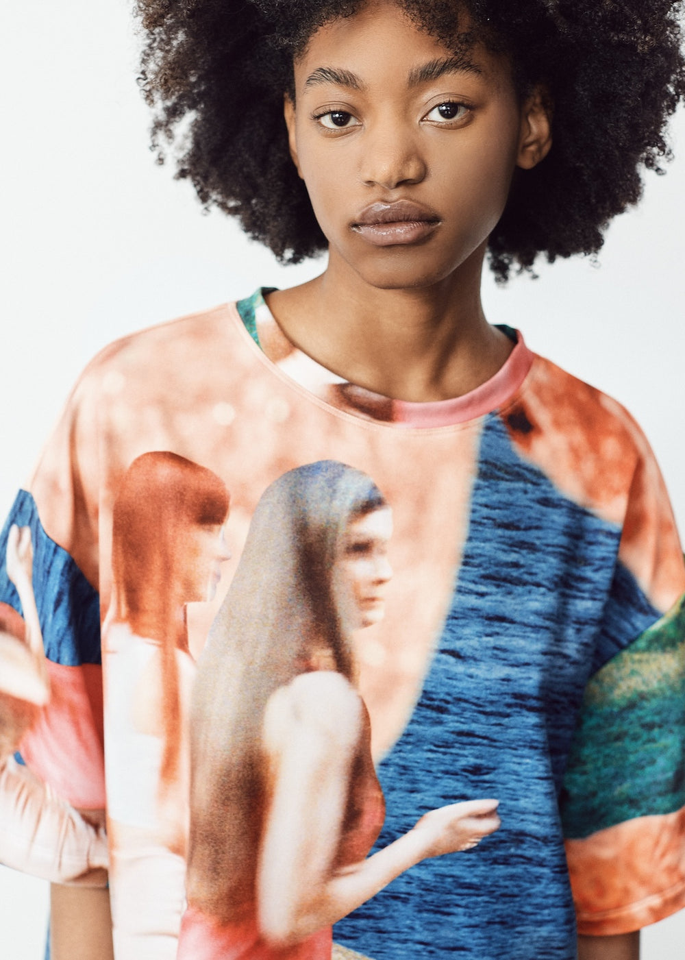 Model in a vibrant, oversized T-shirt with abstract blue, green, and orange patterns, paired with light knee-length shorts featuring black panel accents. The model has natural curly hair and wears flip-flops, standing in a relaxed pose against a minimalistic backdrop