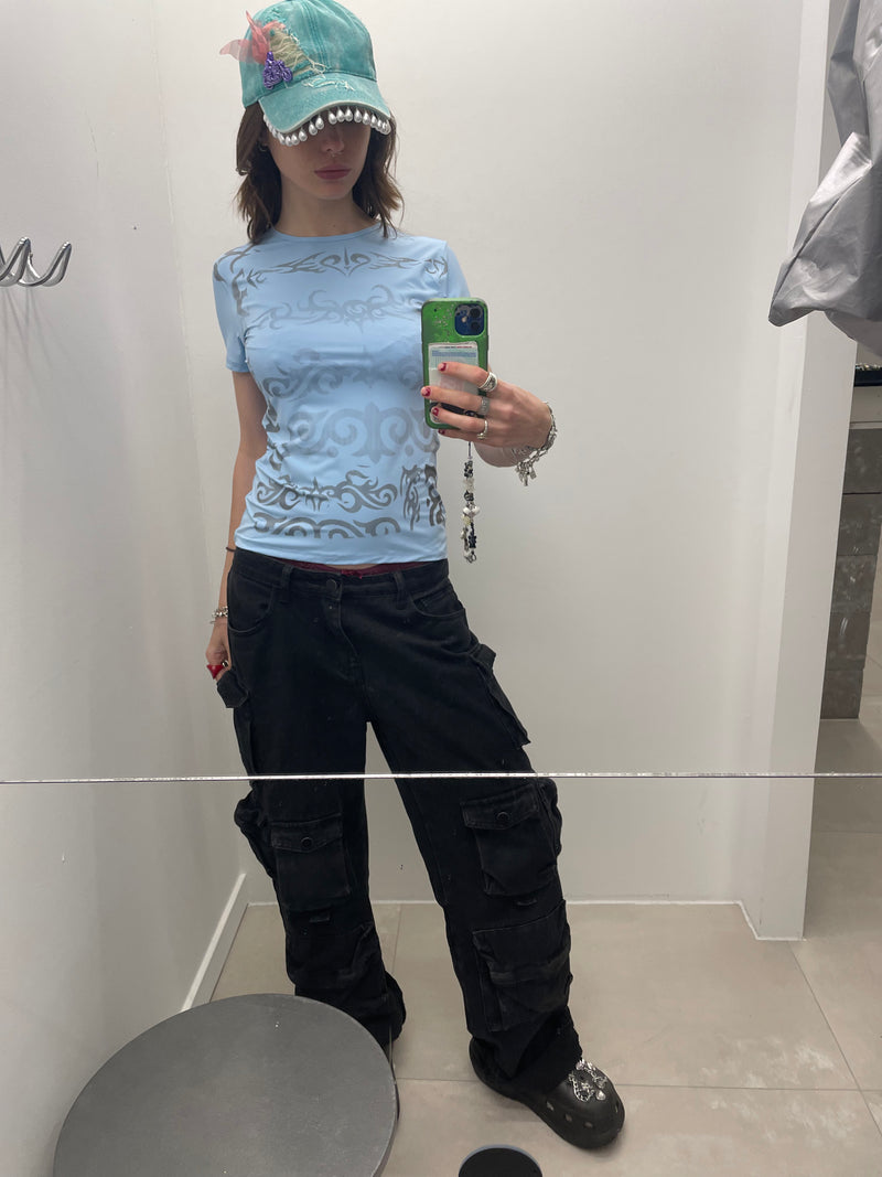 Person taking a mirror selfie wearing a light blue patterned shirt, black cargo pants, and a green cap.