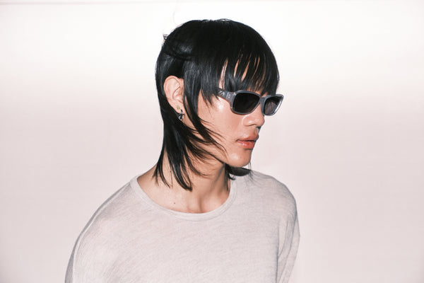 Person with stylish haircut and sunglasses wearing a light gray shirt on a plain background