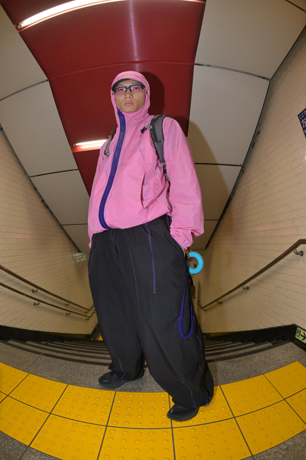 Athletic man standing confidently at the bottom of a subway staircase, dressed in a pink technical hooded jacket and black pants with purple seam accents, holding climbing gear and embodying urban adventure