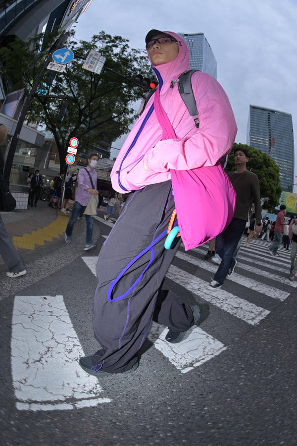 Athletic man crossing a busy urban crosswalk in a bright pink technical jacket and black climbing pants with purple accents, carrying gear in a matching pink sling bag, exuding confidence and urban functionality