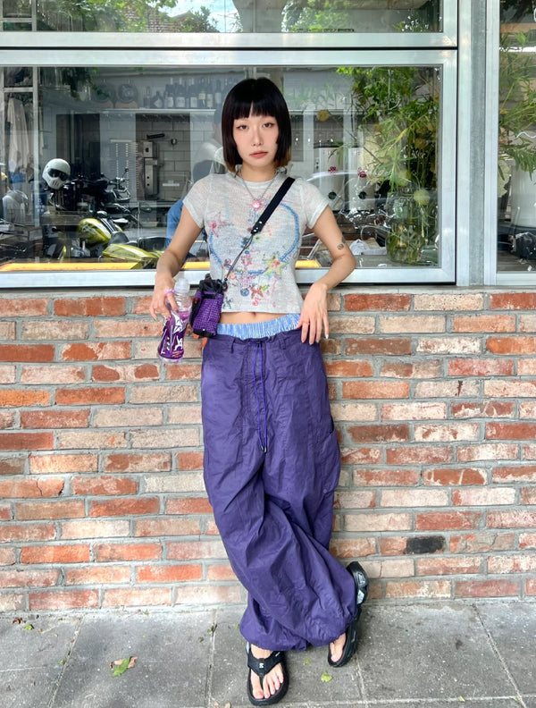 Woman leaning against a brick wall outside a café, wearing loose-fit purple drawstring pants and a sheer floral top, accessorized with a crossbody strap and bottle holder, capturing relaxed urban summer style.