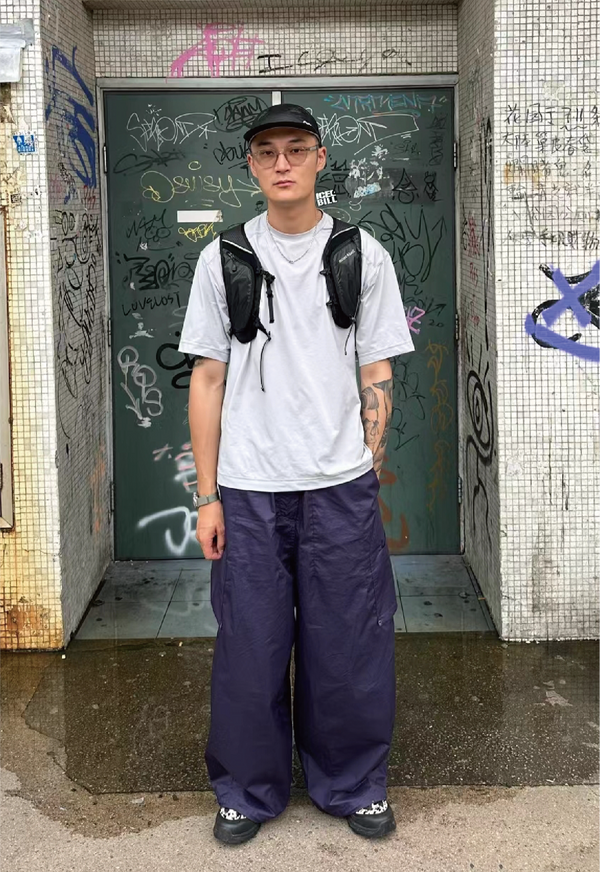 Man standing in front of a graffiti-covered doorway, wearing loose-fit purple cargo pants, a white T-shirt, black cap, and tactical chest rig, showcasing a blend of streetwear and utilitarian fashion.