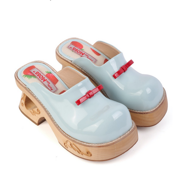 A pair of light blue glossy clogs with wooden platform soles. The shoes feature cut-out details in the heel and are adorned with small red bow accents on the upper. The insole has a strawberry-themed design with the text “Bonne Maison BOSS.” The background is white, highlighting the unique design of the clogs. Keywords, kawaii, strawberry print, very comfy