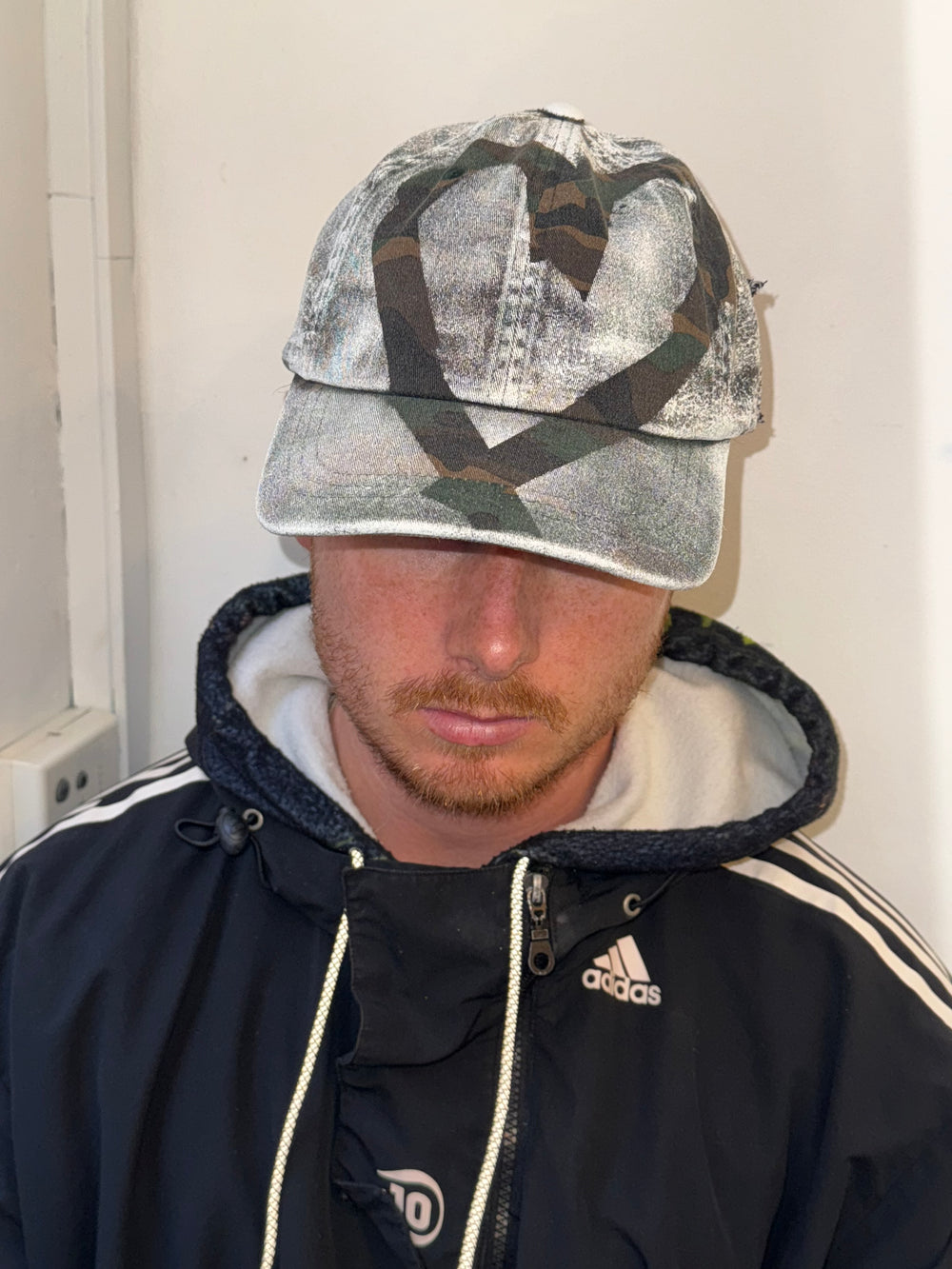 “A distressed camouflage baseball cap featuring a mix of green, brown, and beige tones. The cap has a worn, vintage texture, with a stitched black patch on the side displaying the handwritten text ‘ROBIN ROSBAK’ in a graffiti-like font. The brim is curved, and the cap is styled casually on a person wearing a black Adidas jacket