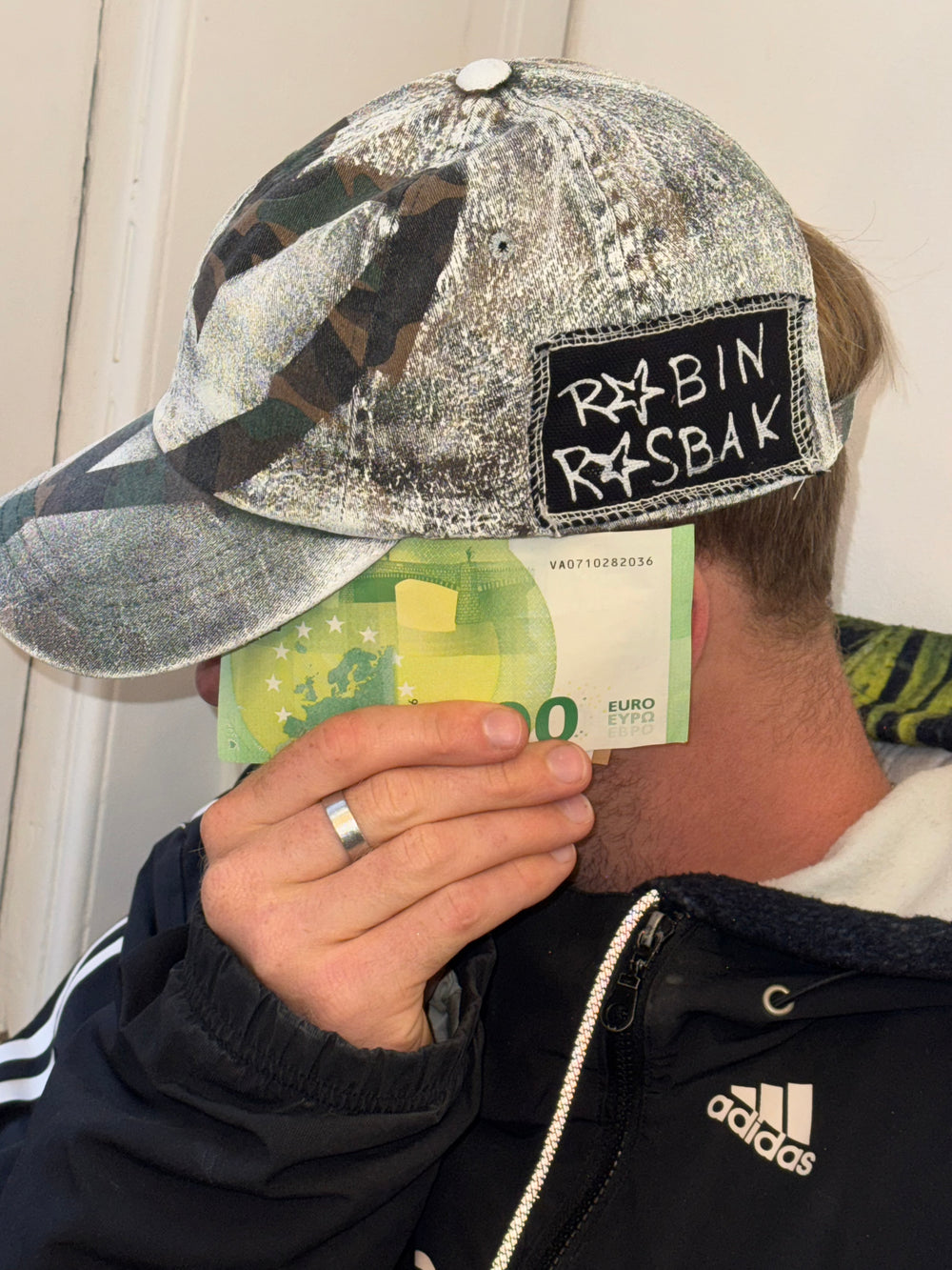 “A distressed camouflage baseball cap featuring a mix of green, brown, and beige tones. The cap has a worn, vintage texture, with a stitched black patch on the side displaying the handwritten text ‘ROBIN ROSBAK’ in a graffiti-like font. The brim is curved, and the cap is styled casually on a person wearing a black Adidas jacket