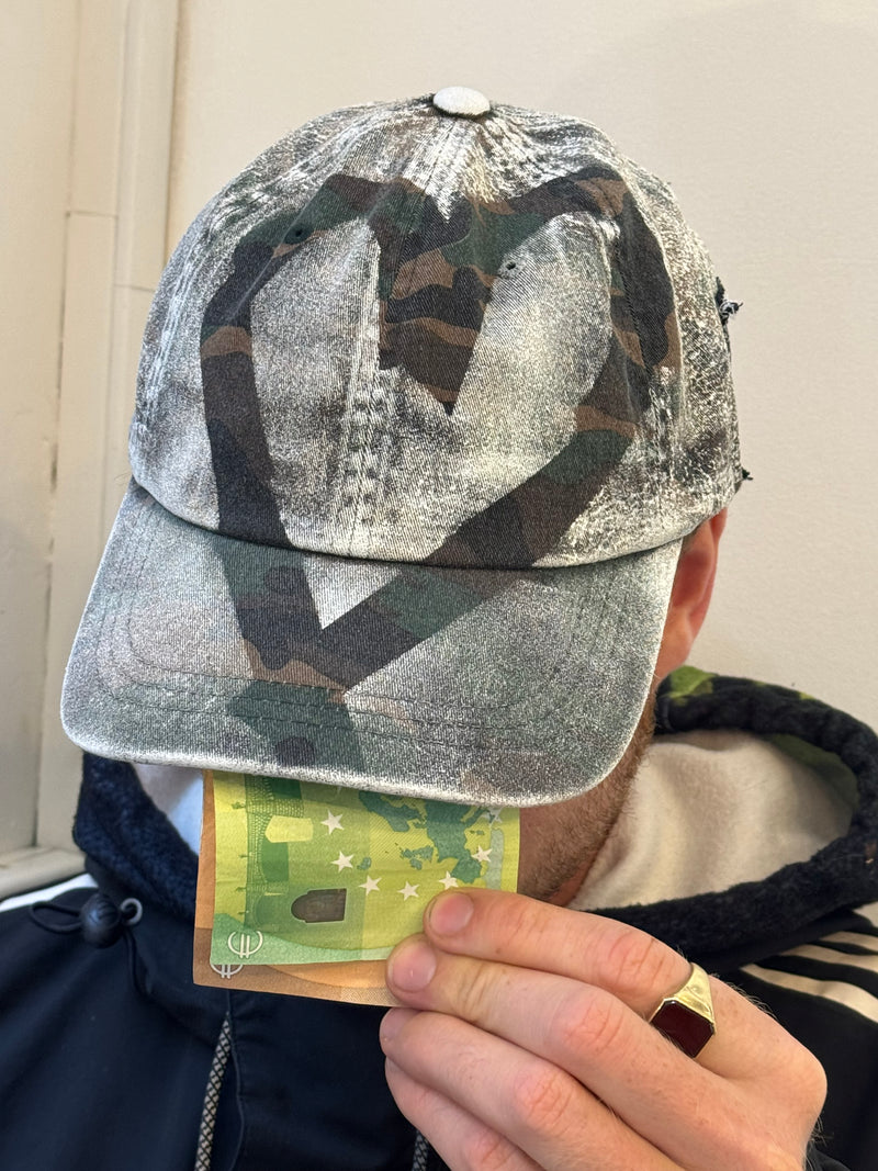 “A distressed camouflage baseball cap featuring a mix of green, brown, and beige tones. The cap has a worn, vintage texture, with a stitched black patch on the side displaying the handwritten text ‘ROBIN ROSBAK’ in a graffiti-like font. The brim is curved, and the cap is styled casually on a person wearing a black Adidas jacket