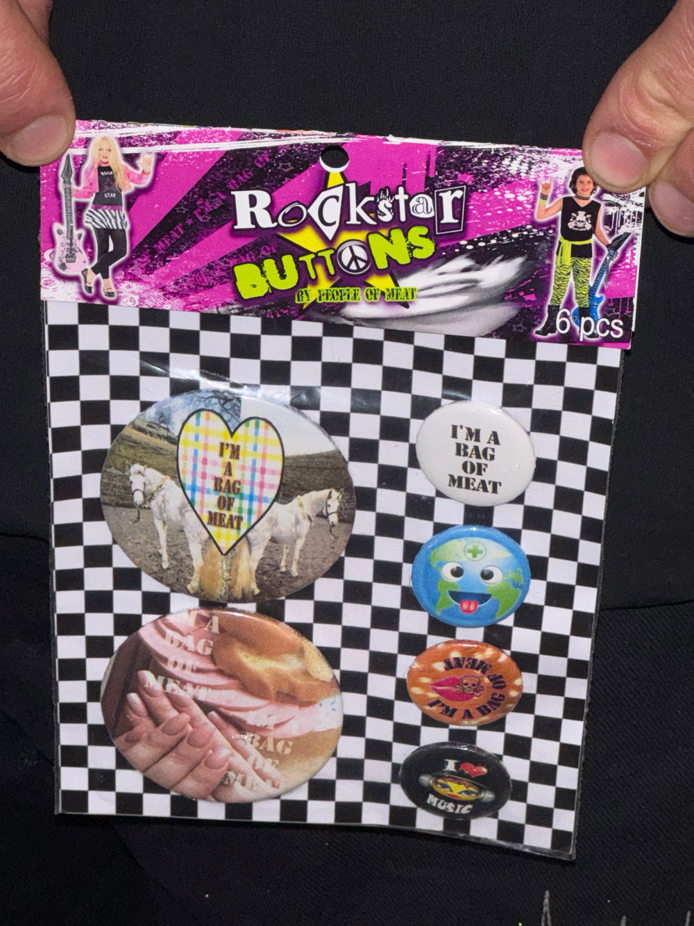 “Packaging for a set of six novelty buttons labeled ‘Rockstar Buttons by People of Meat.’ The top of the package features a vibrant pink and black design with a rocker-themed aesthetic, including images of two people dressed in rockstar outfits holding guitars. The bottom half displays six buttons against a black-and-white checkered background. The buttons include designs such as a heart with ‘I’m a bag of meat,’ an Earth character, and slogans like ‘I love music’ and ‘I’m a bag of meat’ with varied imagery