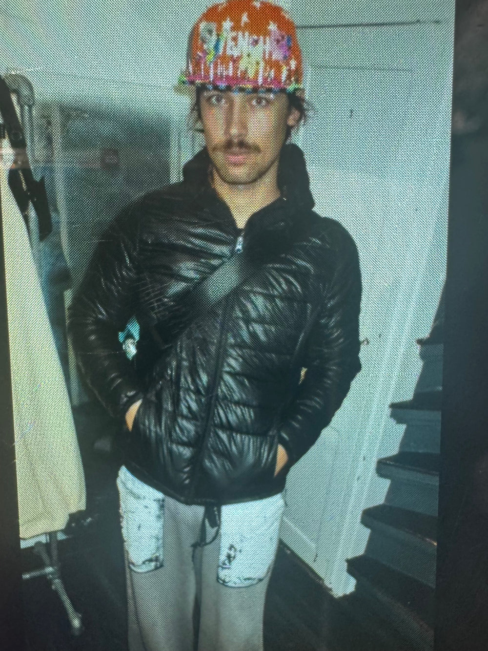 A man standing indoors wearing a black puffy jacket, gray sweatpants with white patches on the knees, and a brightly colored, patterned hat. He has a mustache and a crossbody bag strap over his jacket. The background shows a staircase and clothing rack in a dimly lit room.”
