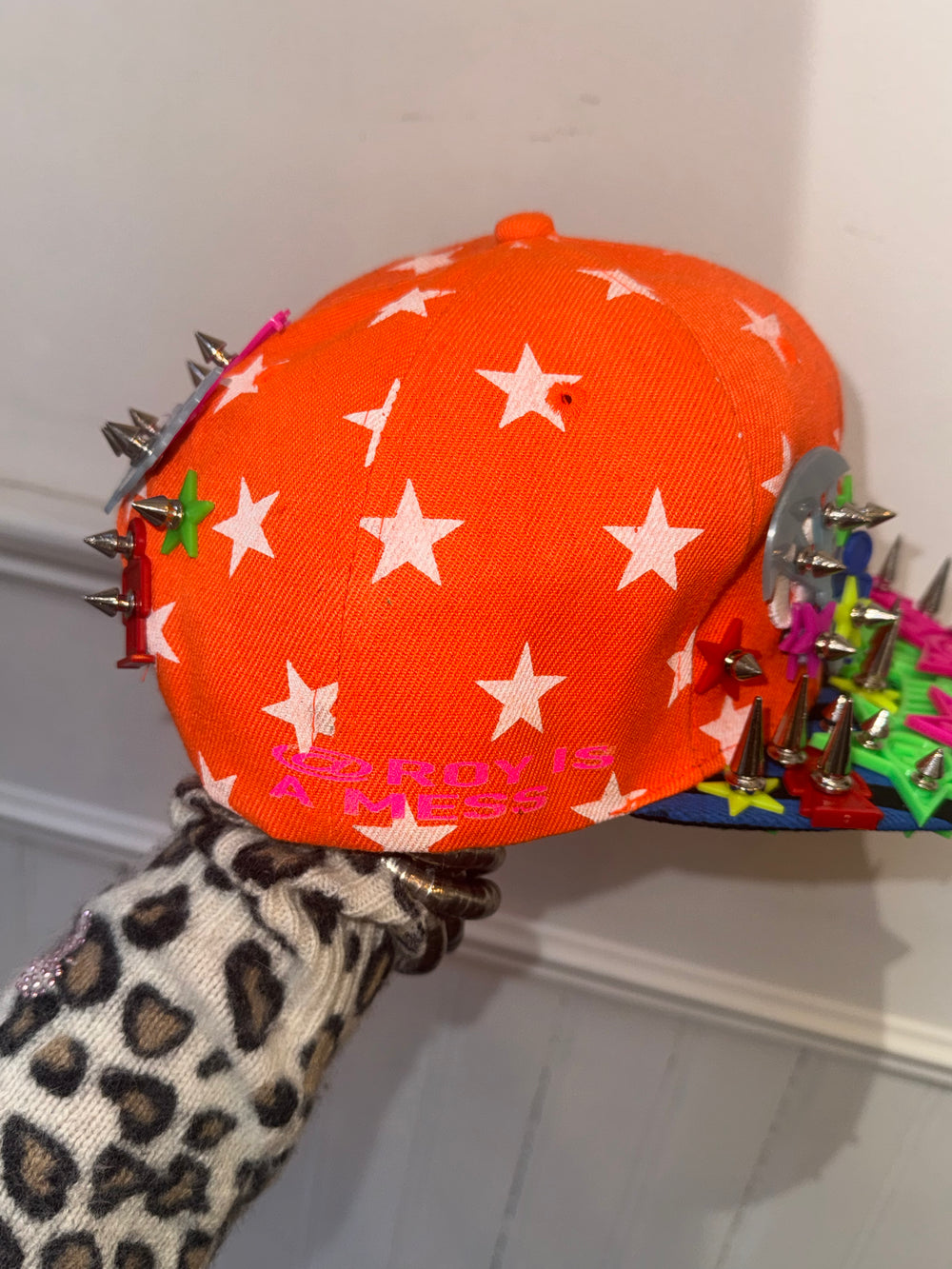 The side view of an orange baseball cap patterned with white stars and adorned with colorful spikes and star-shaped embellishments. The text ‘ROY IS A MESS’ is printed in bold pink letters on the back panel. The cap is being held up by a hand wearing a leopard-print sleeve.