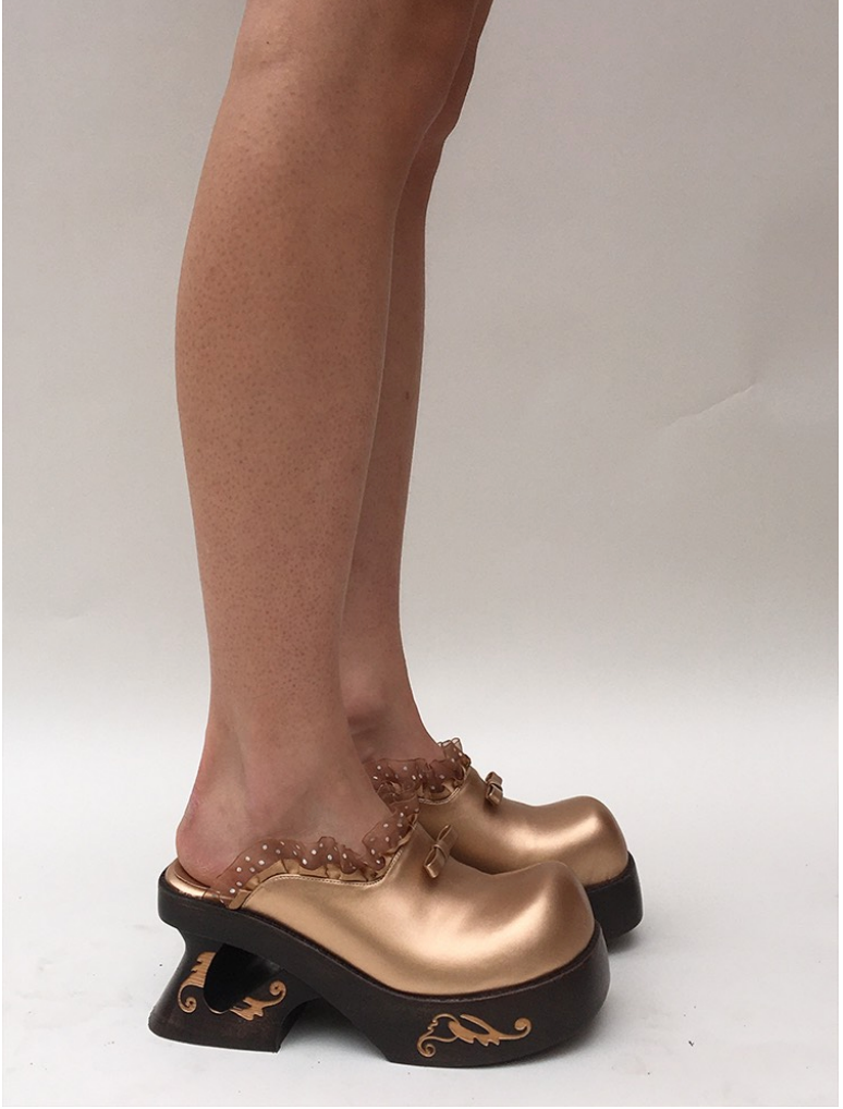 Unique gold-toned clogs with a glossy finish, featuring delicate polka-dotted ruffle trim on the upper and a decorative cut-out heel design. The base is dark with gold filigree patterns, combining whimsical and luxurious elements in their design. The cute gold bow and lace make it a very cute design. Keywords: harajuku, lace ruffle details, cute fashion, bow, grape shoes