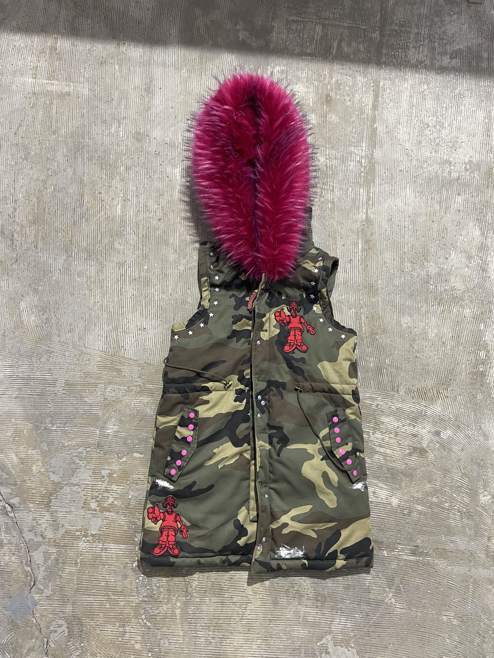 Camouflage hooded padded vest, front view, with snap-button closure, graphic patches, stud details, and pink faux fur hood trim.