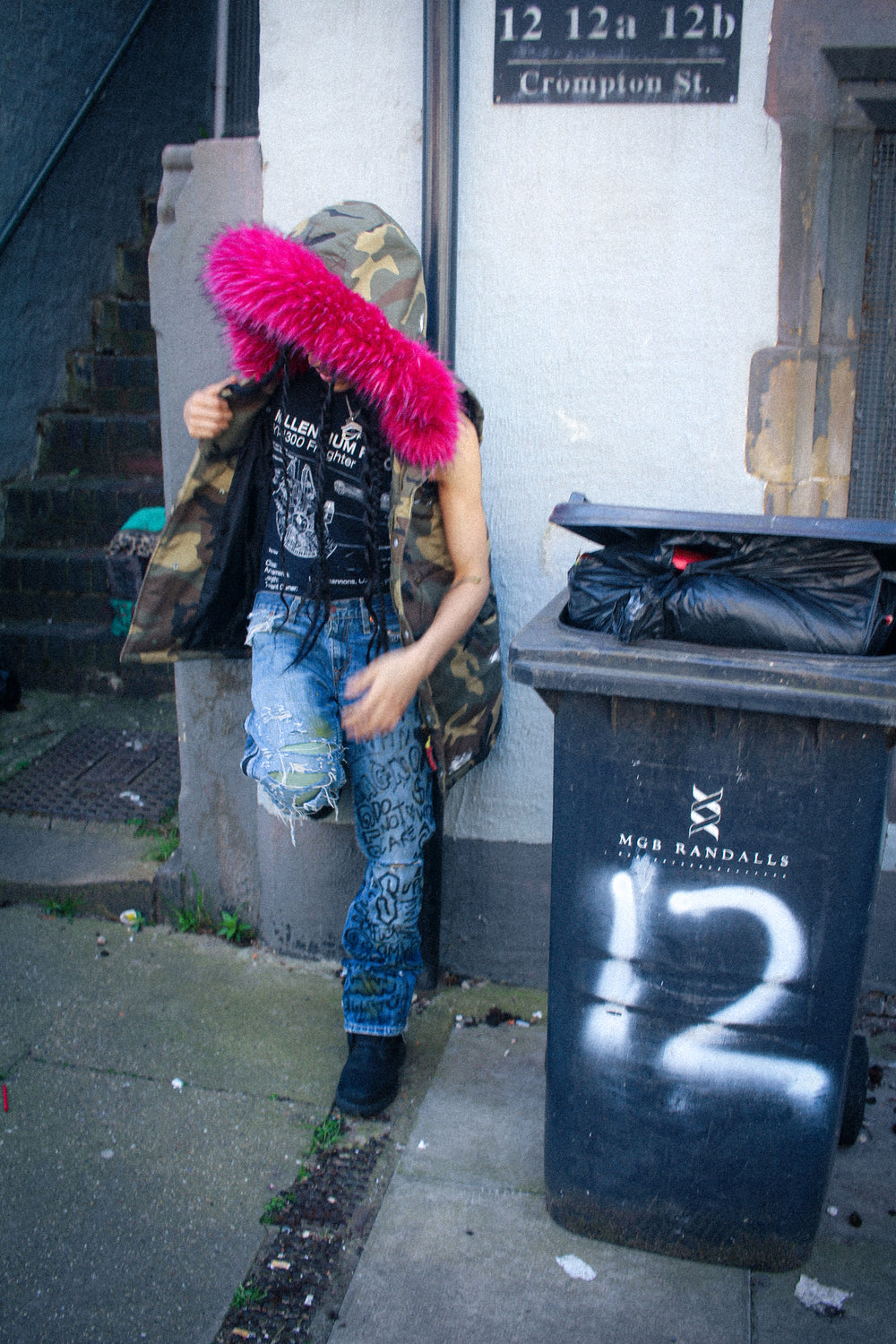 Camouflage hooded padded vest worn outdoors, front view with pink faux fur hood trim and printed graphic patches.