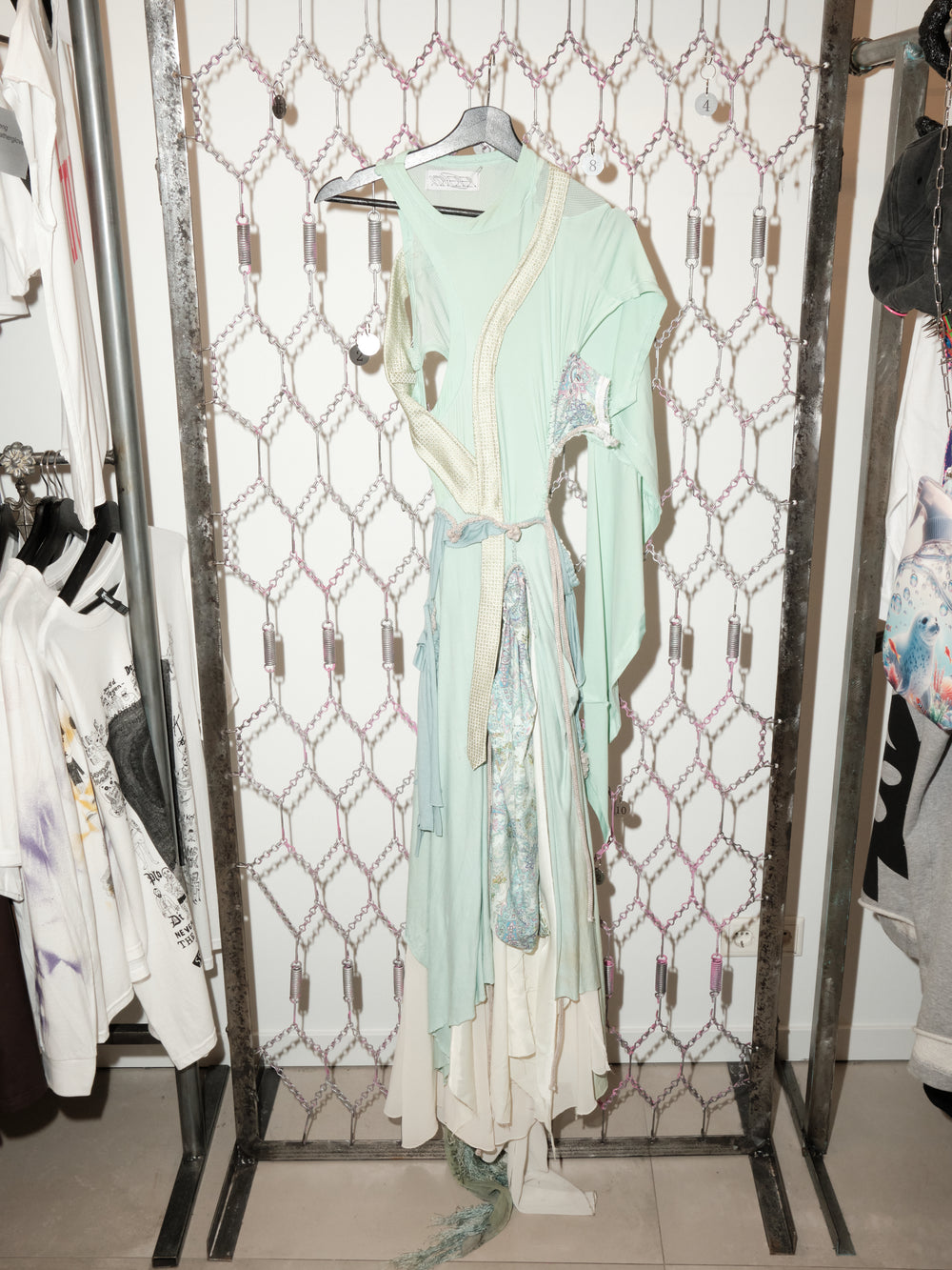 A long, draped dress in pale mint green and cream hangs on a hanger against a chain-link patterned backdrop. The dress features asymmetrical cutouts, layered flowing fabric, and decorative pastel panels with a textured sash tied at the waist. Its trailing hem and mixed materials give it an ethereal, deconstructed look.