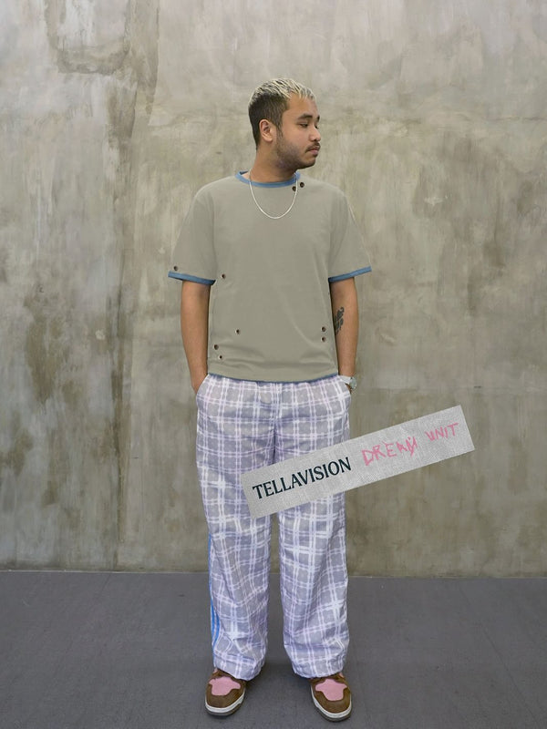 Model wearing Tellavision Eyelet T-Shirt in muted grey with blue trim and metal eyelet details styled with pastel tartan trousers.