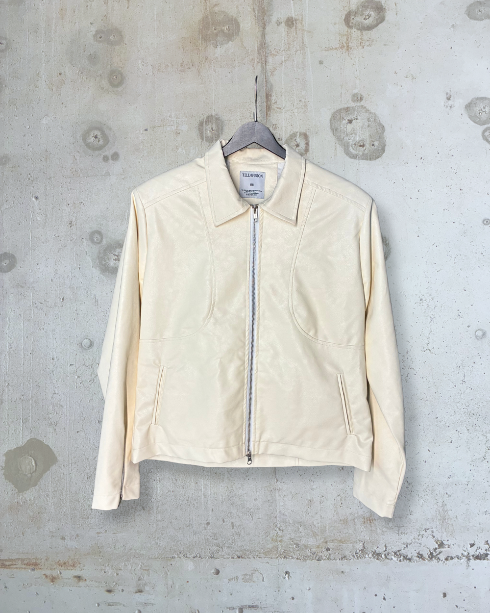 Cream faux leather zip-up jacket by Tellavision with structured seams, pointed collar, and front pockets hanging against concrete wall.