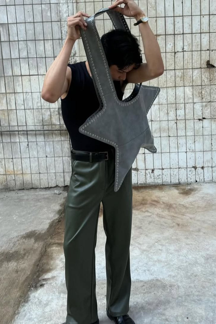 Person holding a large gray star-shaped bag and wearing green faux leather pants  against a tiled wall.