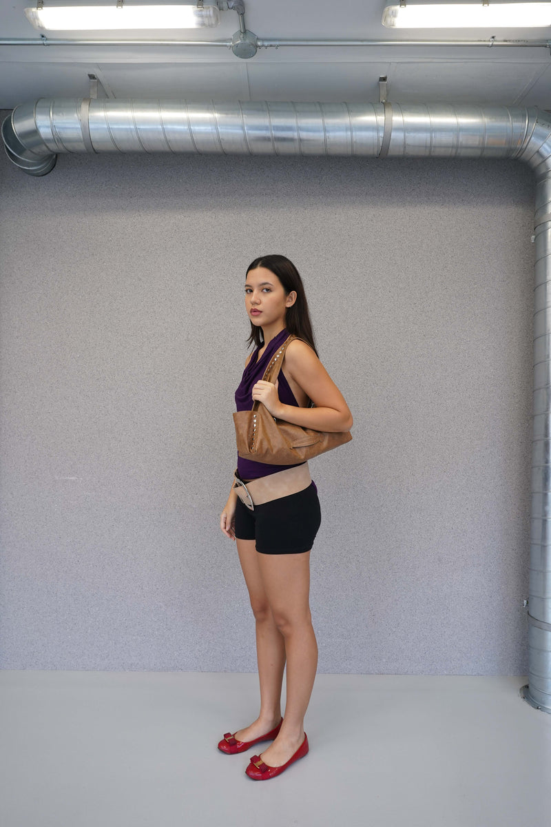 Woman standing in a room with gray walls and visible pipes, holding a brown bag.
