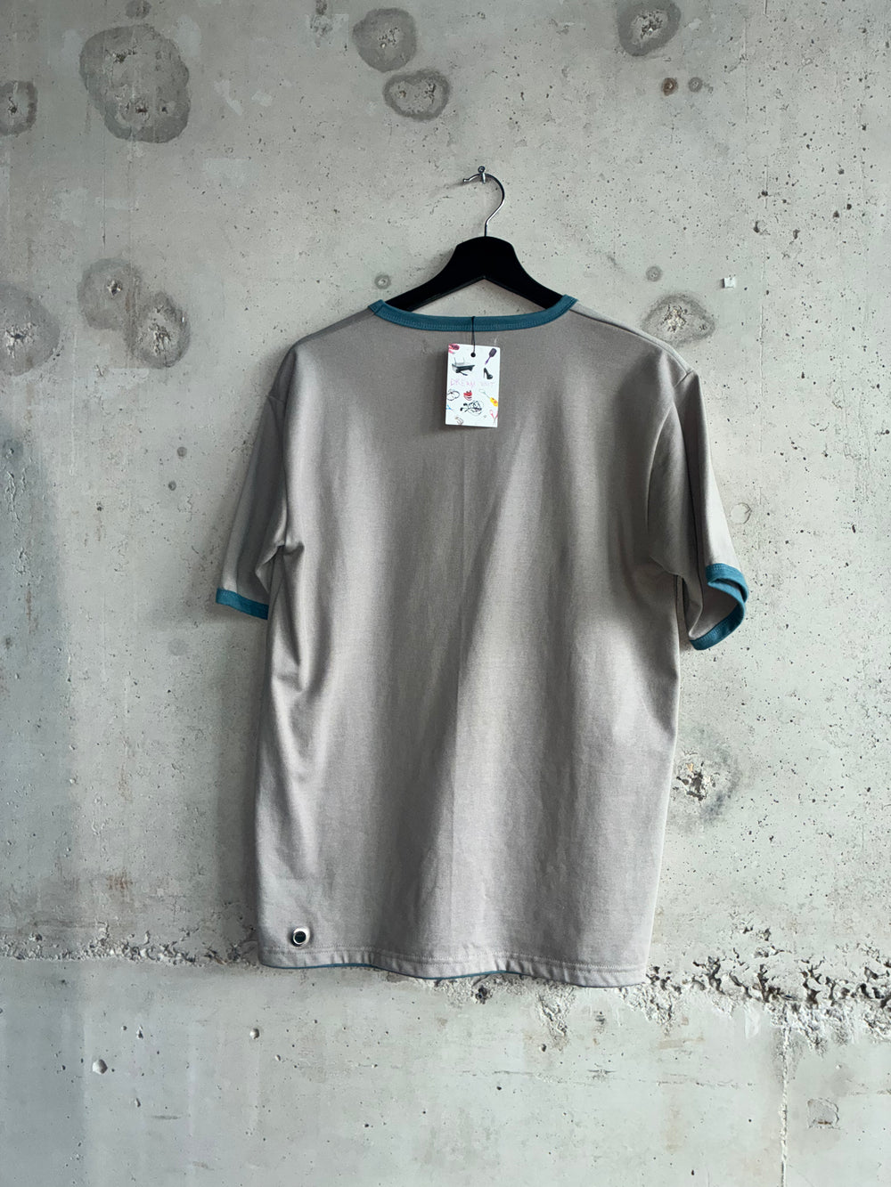 Back view of the Tellavision Eyelet T-shirt in grey with blue trim and a single silver eyelet detail near the hem, hanging against a raw concrete wall.