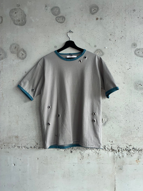 Front view of the Tellavision Eyelet T-shirt in grey with blue trim and silver eyelet hardware details, displayed against a textured concrete wall.