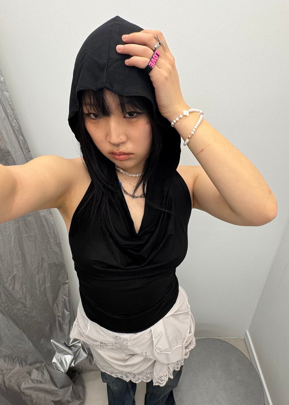 High-angle selfie of a black hooded cowl neck top paired with a white lace-trimmed tiered skirt and baggy jeans. Edgy streetwear style with chunky rings and bead bracelets.