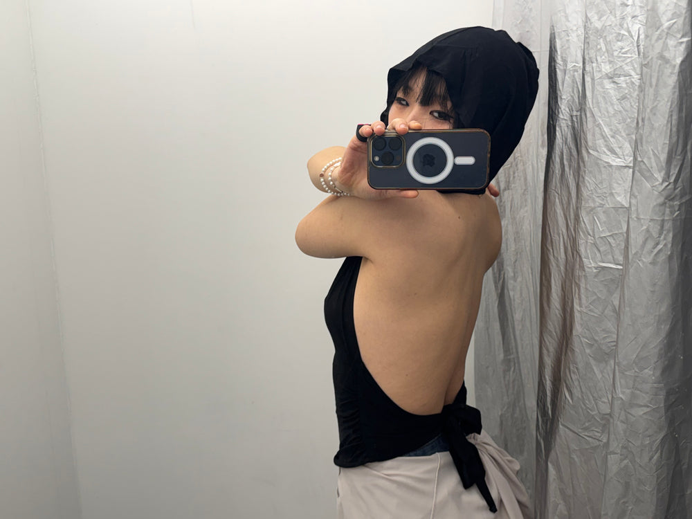 Side profile of a black backless hooded top featuring a wrap-around tie closure at the waist. Styled with a neutral-toned layered skirt for a "grunge-sleek" aesthetic.