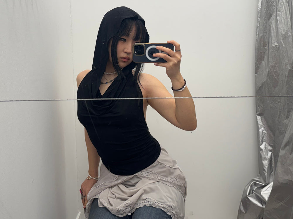 Mirror selfie featuring a black sleeveless hooded top with a soft cowl neckline. Paired with a lace-edge mini skirt over jeans for a layered 2000s-inspired outfit.